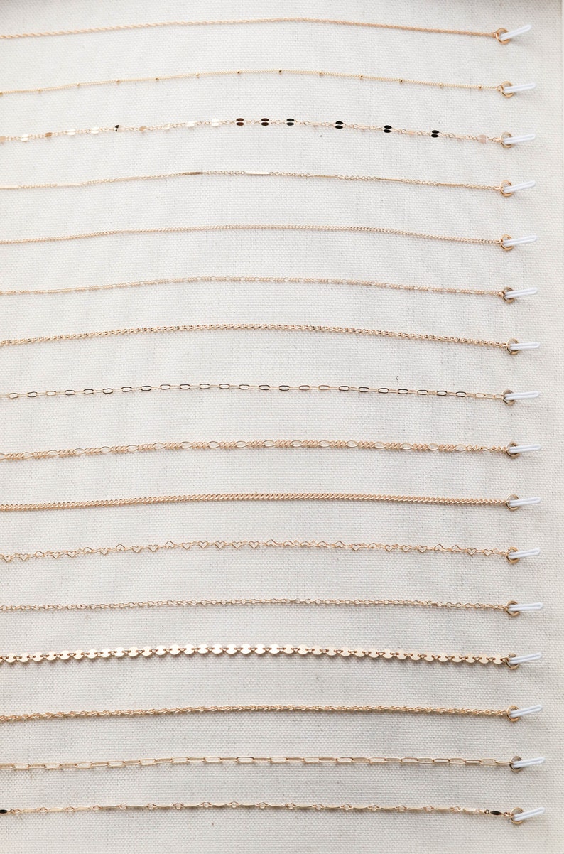 May include: A display of 14 different gold chains, each with a unique link style, hanging from white hooks on a beige fabric background.