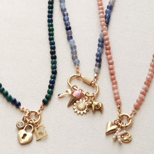 May include: Three beaded necklaces with gold-colored clasps and charms. One necklace has green and blue beads, a heart-shaped lock charm, and an "E" charm. Another has blue and pink beads, and charms including a key, ice cream cone, sunflower, and elephant.
