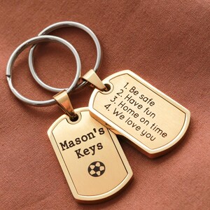 May include: Two gold-toned metal dog tag keychains attached to silver-colored split rings. One tag is engraved with 'Mason's Keys' and a soccer ball icon. The other tag features the text: '1. Be safe, 2. Have fun, 3. Home on time, 4. We love you'.