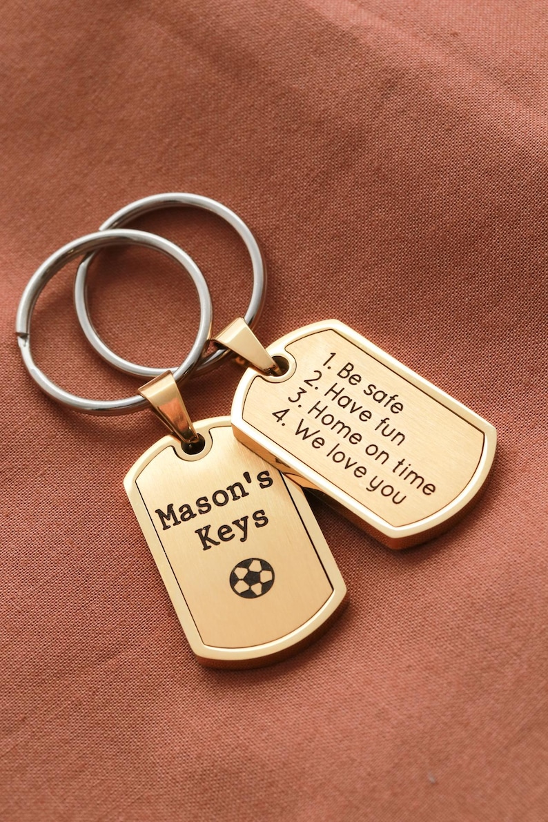 May include: Two gold-toned metal dog tag keychains attached to silver-colored split rings. One tag is engraved with 'Mason's Keys' and a soccer ball icon. The other tag features the text: '1. Be safe, 2. Have fun, 3. Home on time, 4. We love you'.