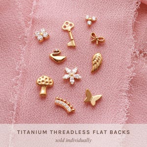 May include: A collection of gold-colored titanium threadless flat backs in various shapes, including a key, butterfly, bow, and mushroom, displayed on a pink fabric. Some pieces are adorned with clear gemstones. The text "TITANIUM THREADLESS FLAT BACKS" is visible.