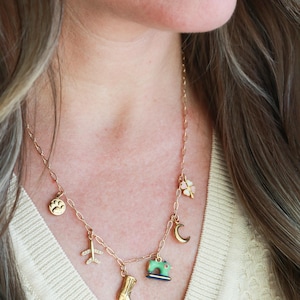 May include: A gold-colored necklace with multiple charms, including a paw print, airplane, cowboy boot, sewing machine, four-leaf clover, and crescent moon. The necklace is worn around the neck and is a delicate piece of jewelry.