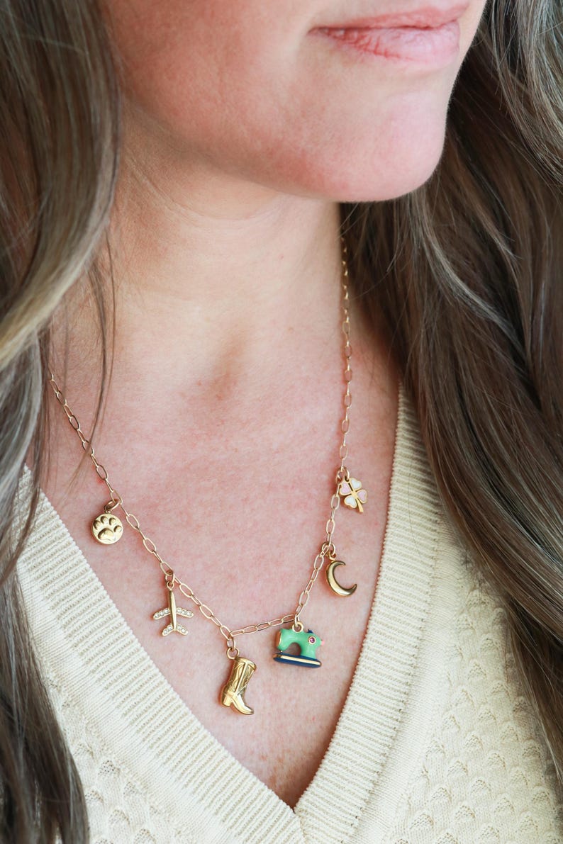 May include: A gold-colored necklace with multiple charms, including a paw print, airplane, cowboy boot, sewing machine, four-leaf clover, and crescent moon. The necklace is worn around the neck and is a delicate piece of jewelry.