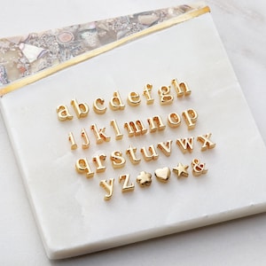 May include: Gold-colored alphabet letter charms and symbols on a white background. The letters include a, b, c, d, e, f, g, h, i, j, k, l, m, n, o, p, q, r, s, t, u, v, w, x, y, z. The symbols include a four-leaf clover, a heart, a star, and an ampersand.