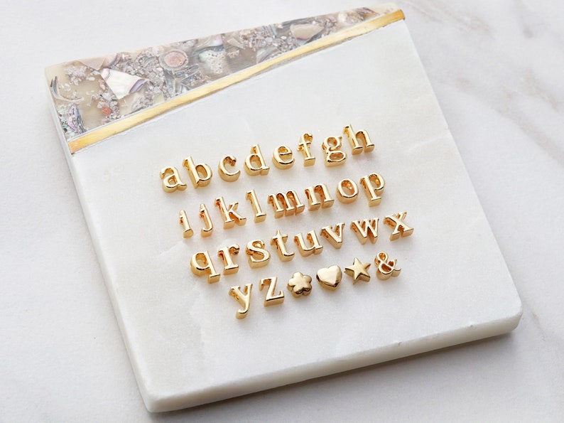 May include: Gold-colored alphabet letter charms and symbols on a white background. The letters include a, b, c, d, e, f, g, h, i, j, k, l, m, n, o, p, q, r, s, t, u, v, w, x, y, z. The symbols include a four-leaf clover, a heart, a star, and an ampersand.