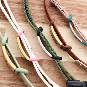 May include: A selection of colourful eyeglass straps. The straps are in various colours, including green, white, brown, and pink. Each strap has a metal accent piece with text. The straps are on a wooden surface.