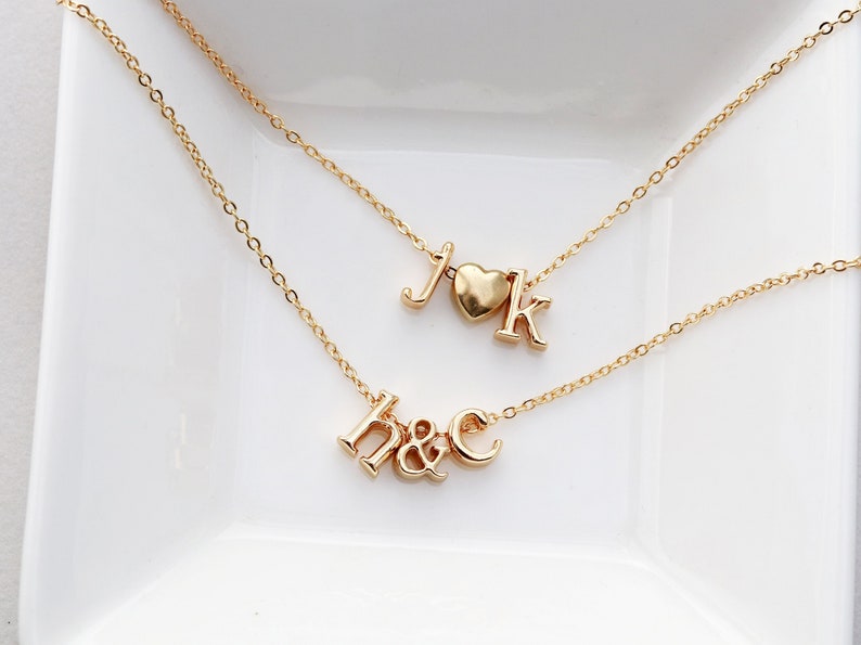 May include: Two gold necklaces with initial charms. One necklace has the letters J and K with a heart charm between them. The other necklace has the letters H and C with an ampersand between them.
