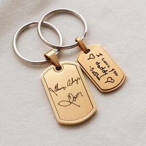 May include: Two gold-toned metal dog tag keychains with silver-toned split rings. The larger tag features a cursive signature, while the smaller tag is engraved with the text I Love you daddy -Sasha and two heart symbols in black ink.