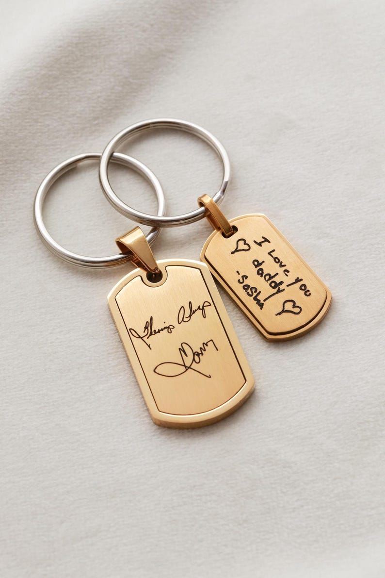 May include: Two gold-toned metal dog tag keychains with silver-toned split rings. The larger tag features a cursive signature, while the smaller tag is engraved with the text I Love you daddy -Sasha and two heart symbols in black ink.