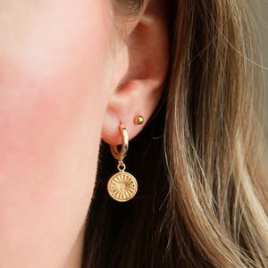 May include: Gold hoop earrings with a small, round, gold charm hanging from one earring. The charm has a detailed, sun-like design.