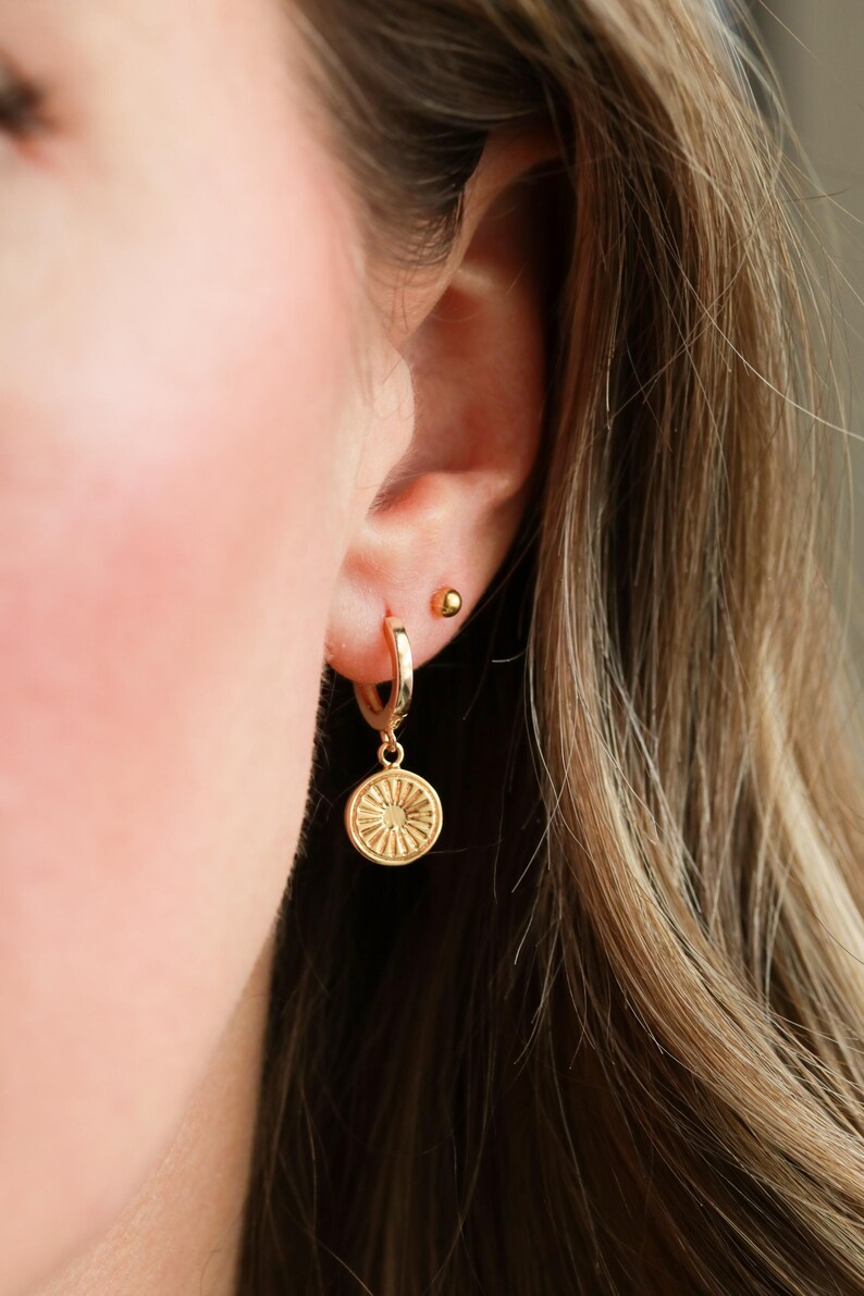 May include: Gold hoop earrings with a small, round, gold charm hanging from one earring. The charm has a detailed, sun-like design.