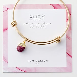May include: A gold bangle bracelet with a ruby gemstone charm. The bracelet is on a white card with the text "RUBY natural gemstone collection TOM DESIGN SHOP".