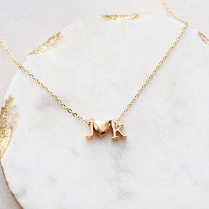 May include: A gold chain necklace with a heart charm and the letters "J" and "K" hanging from it. The necklace is on a white marble surface.