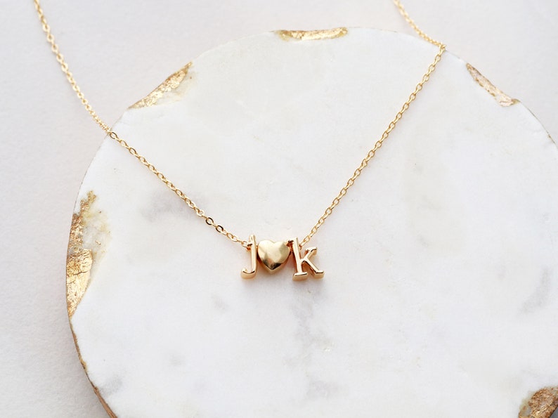 May include: A gold chain necklace with a heart charm and the letters "J" and "K" hanging from it. The necklace is on a white marble surface.