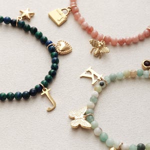May include: Close-up of four beaded bracelets with gold charms. One bracelet has dark blue and green beads with a star, heart, and letter "J" charm. Another has pink beads with a bee and letter "S" charm. The other two have a strawberry, lock, and butterfly charm.