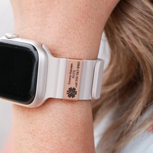 May include: A silver smartwatch with a light gray silicone band featuring a rose gold-colored metal medical alert tag. The tag is engraved with the text Emma Hansen, POTS, call 555-567-8901, and a black medical star of life symbol.