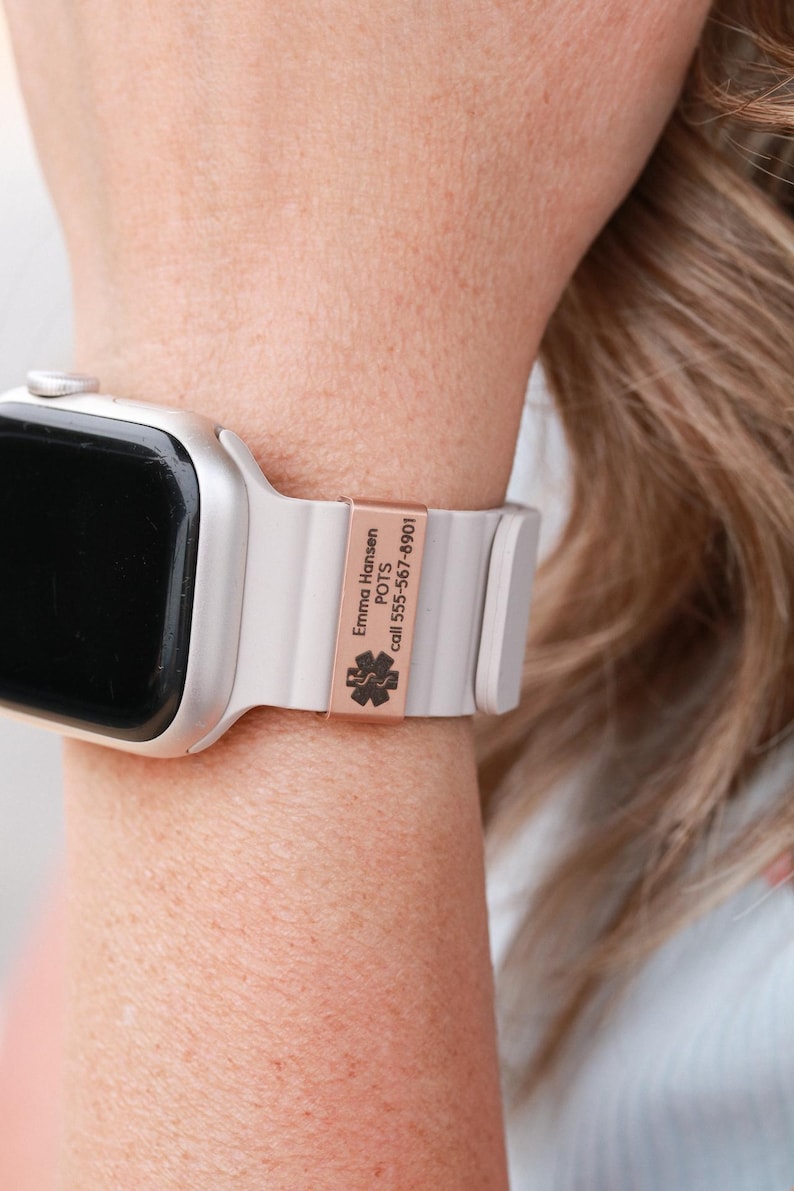 May include: A silver smartwatch with a light gray silicone band featuring a rose gold-colored metal medical alert tag. The tag is engraved with the text Emma Hansen, POTS, call 555-567-8901, and a black medical star of life symbol.