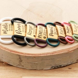 May include: A selection of personalised pet ID tags, each with a name and phone number. The tags have gold-coloured metal plates with different names and matching coloured silicone loops. They are displayed on a wooden surface.