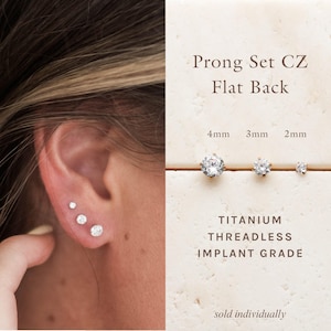 May include: Three prong set cubic zirconia flat back earrings in 4mm, 3mm, and 2mm sizes. The text reads: Prong Set CZ Flat Back, 4mm 3mm 2mm, TITANIUM THREADLESS IMPLANT GRADE, sold individually. These clear stone studs are shown worn in an earlobe.