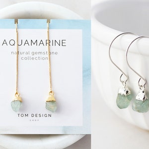 May include: A pair of aquamarine gemstone earrings with gold-plated chains and silver hooks. The earrings are displayed on a white card with the text "AQUAMARINE natural gemstone collection TOM DESIGN SHOP".