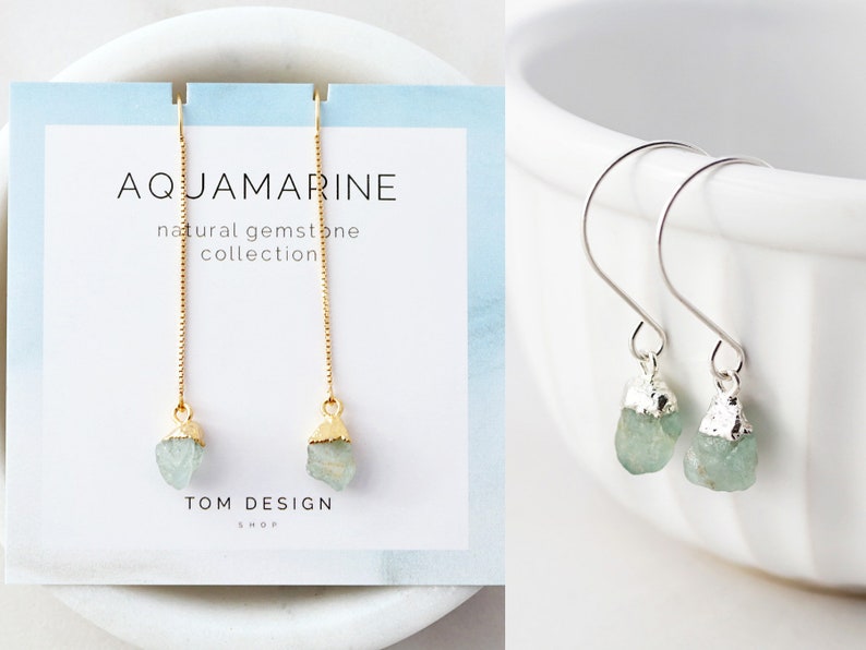 May include: A pair of aquamarine gemstone earrings with gold-plated chains and silver hooks. The earrings are displayed on a white card with the text "AQUAMARINE natural gemstone collection TOM DESIGN SHOP".