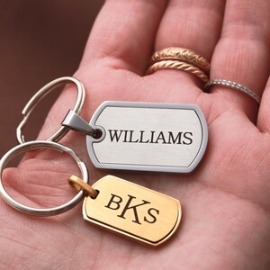 May include: Two rectangular metal dog tag keychains with rounded corners. One tag is silver-toned with the engraved text WILLIAMS, and the other is gold-toned with the engraved text BKS. Both tags are attached to circular metal split key rings.