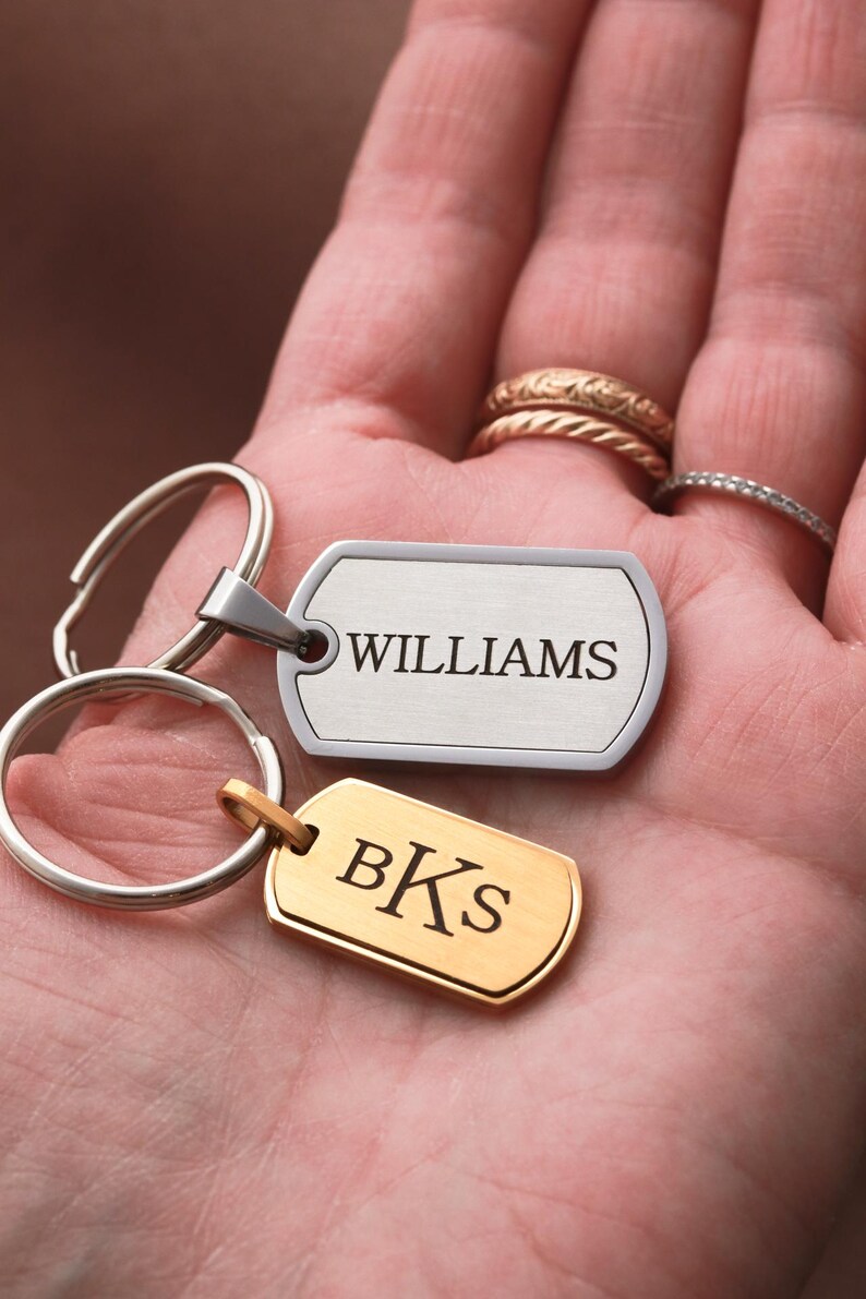 May include: Two rectangular metal dog tag keychains with rounded corners. One tag is silver-toned with the engraved text WILLIAMS, and the other is gold-toned with the engraved text BKS. Both tags are attached to circular metal split key rings.