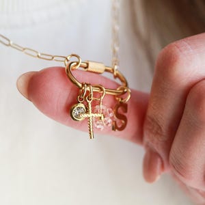 May include: Gold-colored necklace with a carabiner clasp and multiple charms. Charms include a cross, a crystal, and the letter "S". The necklace is held by a finger, showcasing the delicate design and craftsmanship.
