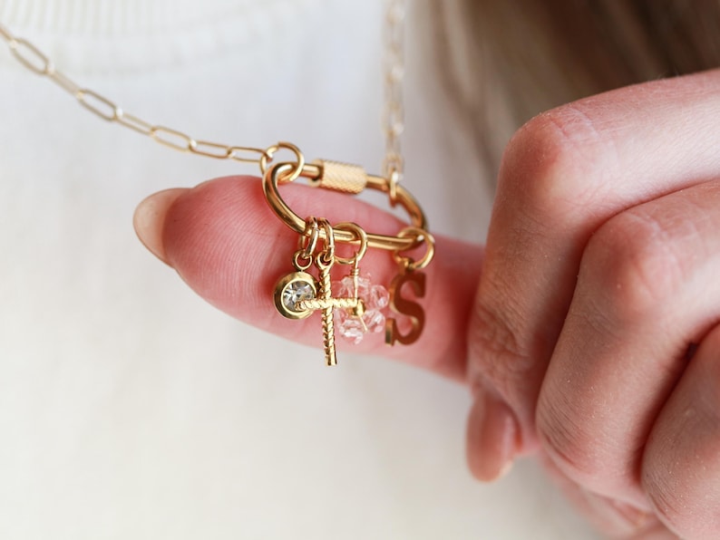 May include: Gold-colored necklace with a carabiner clasp and multiple charms. Charms include a cross, a crystal, and the letter "S". The necklace is held by a finger, showcasing the delicate design and craftsmanship.