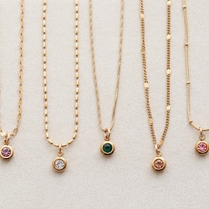 Op de afbeelding: Vijf goudkleurige kettingen met verschillende kettingstijlen. Elke ketting heeft een kleine, ronde hanger met een gekleurde edelsteen: roze, helder, groen, perzik en roze. De hangers zijn gezet in gouden bezels.