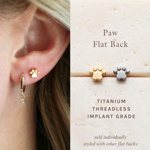 May include: Gold-colored paw print stud earrings and a gold hoop earring with a dangling charm are shown on an ear. The text "Paw Flat Back" is visible. The image also shows a silver paw print stud earring.