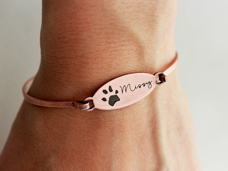 May include: A copper-toned metal bangle bracelet featuring a central oval-shaped charm. The charm is engraved with a black paw print graphic and the name Missy in a cursive script font. The bracelet has a thin, smooth band design.
