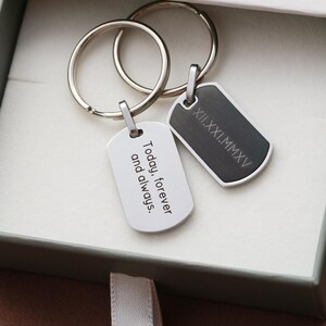 May include: Two metal keychains with rectangular tags attached to silver split rings. One tag is silver with the engraved text Today, forever and always. The other tag is dark gray with the engraved Roman numeral date XII.XXI.MMXV.