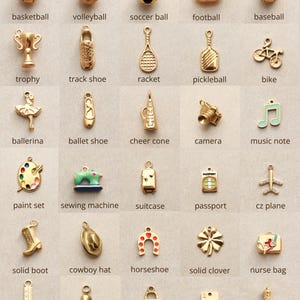 May include: A collection of gold-toned charms in various shapes, including sports equipment like a basketball and a soccer ball, along with other items such as a camera, a sewing machine, and a nurse bag. Each charm has a small loop for attaching to a bracelet or necklace.