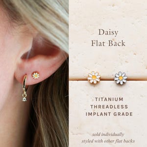 May include: Daisy flat back earrings with white petals and a yellow center. One earring is worn in an ear piercing, and another is displayed next to a matching earring. The earrings are made of titanium.