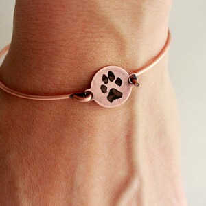 May include: A copper-toned wire bangle bracelet featuring a circular charm with a dark, engraved paw print design. The bracelet has a simple hook-and-eye closure mechanism, and the metal has a warm, metallic finish throughout the entire piece.