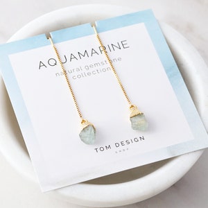 May include: Aquamarine gemstone earrings with gold chain and a light blue gemstone. The earrings are on a white card with the text "Aquamarine natural gemstone collection" and "TOM DESIGN SHOP".