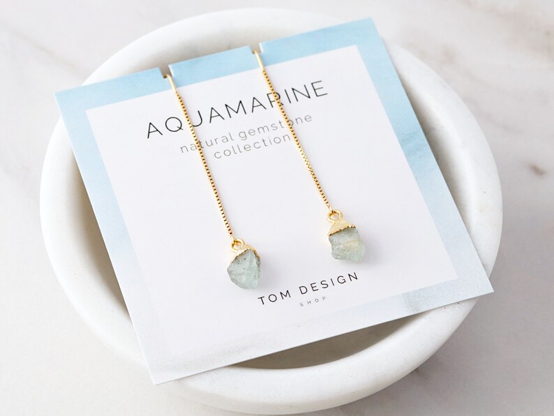 May include: Aquamarine gemstone earrings with gold chain and a light blue gemstone. The earrings are on a white card with the text "Aquamarine natural gemstone collection" and "TOM DESIGN SHOP".
