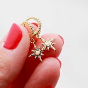 May include: Gold hoop earrings with a dangling star charm featuring a white opal stone and clear cubic zirconia accents.