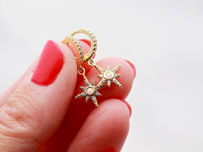 May include: Gold hoop earrings with a dangling star charm featuring a white opal stone and clear cubic zirconia accents.