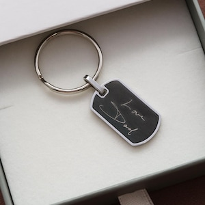 May include: A silver-toned metal dog tag keychain featuring a black rectangular center with the engraved white cursive text Love Dad. The keychain includes a circular split ring attached to the top of the tag, presented inside a white gift box.