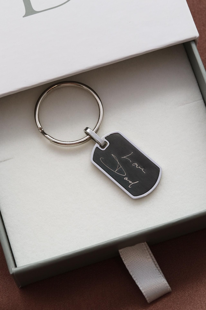 May include: A silver-toned metal dog tag keychain featuring a black rectangular center with the engraved white cursive text Love Dad. The keychain includes a circular split ring attached to the top of the tag, presented inside a white gift box.