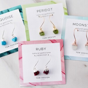 May include: Five cards with different colored backgrounds and silver, gold, and rose gold earring displays. Each card features a different gemstone: turquoise, peridot, moonstone, ruby. The cards all have the text "natural gemstone collection" and "TOM DESIGN" on them.