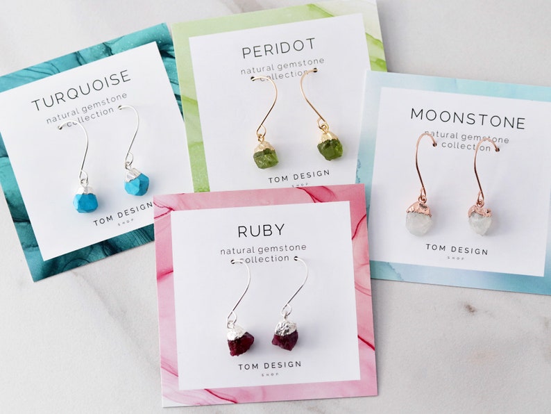 May include: Five cards with different colored backgrounds and silver, gold, and rose gold earring displays. Each card features a different gemstone: turquoise, peridot, moonstone, ruby. The cards all have the text "natural gemstone collection" and "TOM DESIGN" on them.