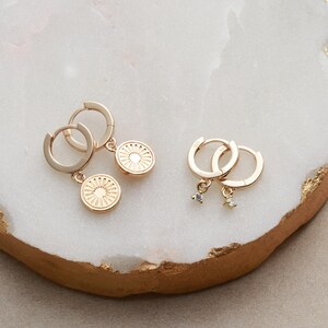May include: Two pairs of gold hoop earrings with charms. One pair has a small gold disc charm with a sun design. The other pair has a small clear crystal charm.