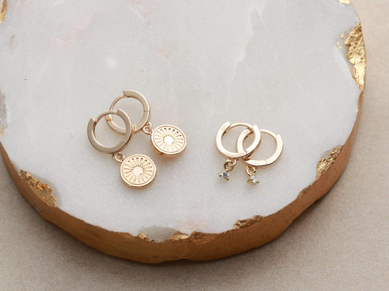 May include: Two pairs of gold hoop earrings with charms. One pair has a small gold disc charm with a sun design. The other pair has a small clear crystal charm.
