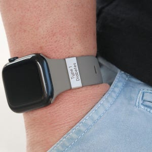 May include: A black smartwatch with a grey silicone band featuring a rectangular silver-toned metal charm engraved with the text Type 1 Diabetes. The watch is worn on a wrist, and the band has a smooth, matte finish with a secure metal loop.