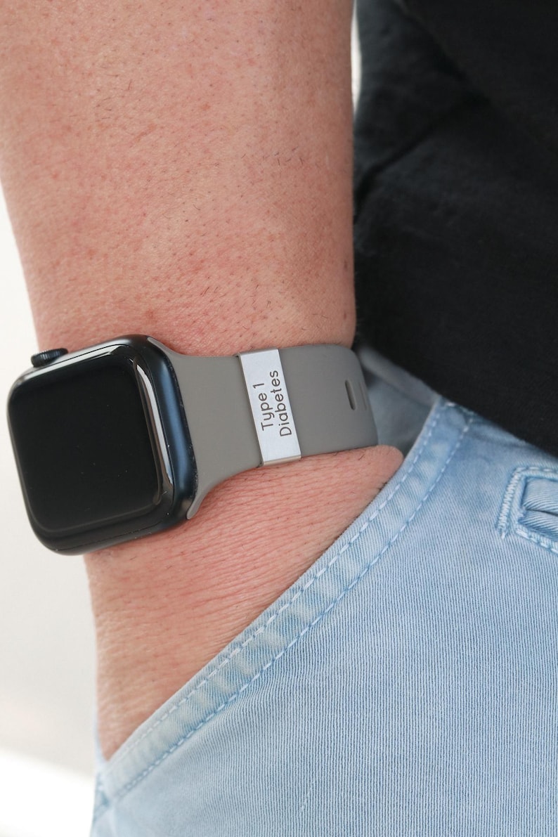 May include: A black smartwatch with a grey silicone band featuring a rectangular silver-toned metal charm engraved with the text Type 1 Diabetes. The watch is worn on a wrist, and the band has a smooth, matte finish with a secure metal loop.