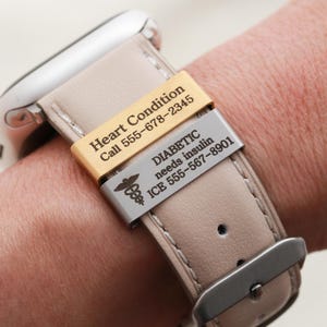 May include: A beige leather watch strap featuring two rectangular metal medical alert tags. The gold tag reads Heart Condition Call 555-678-2345. The silver tag features a caduceus symbol and reads DIABETIC needs insulin ICE 555-567-8901.