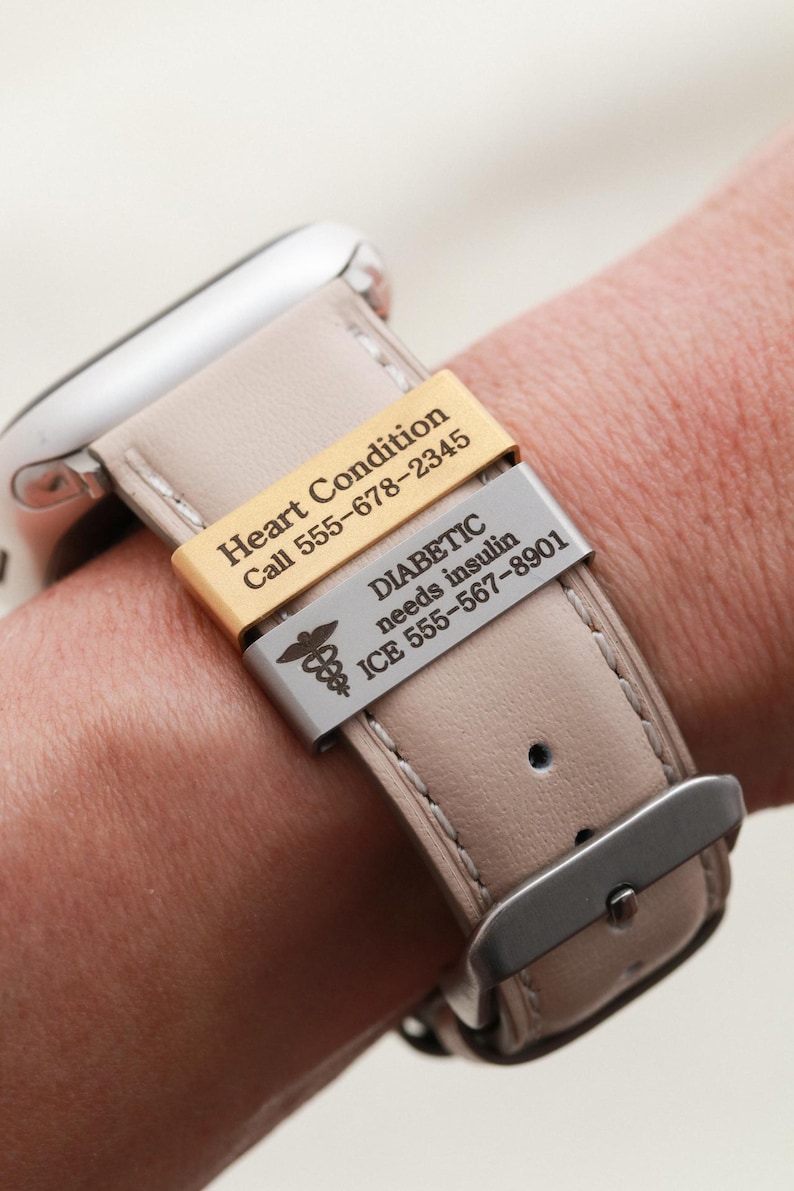 May include: A beige leather watch strap featuring two rectangular metal medical alert tags. The gold tag reads Heart Condition Call 555-678-2345. The silver tag features a caduceus symbol and reads DIABETIC needs insulin ICE 555-567-8901.